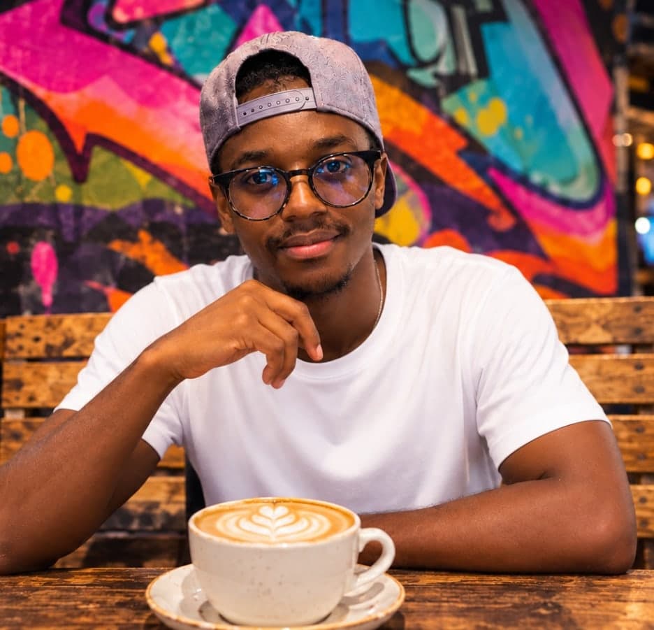 David Eshiwani in a white T-shirt, glasses and a backwards cap, sitting at a café table with a latte in front of a colourful graffiti wall.